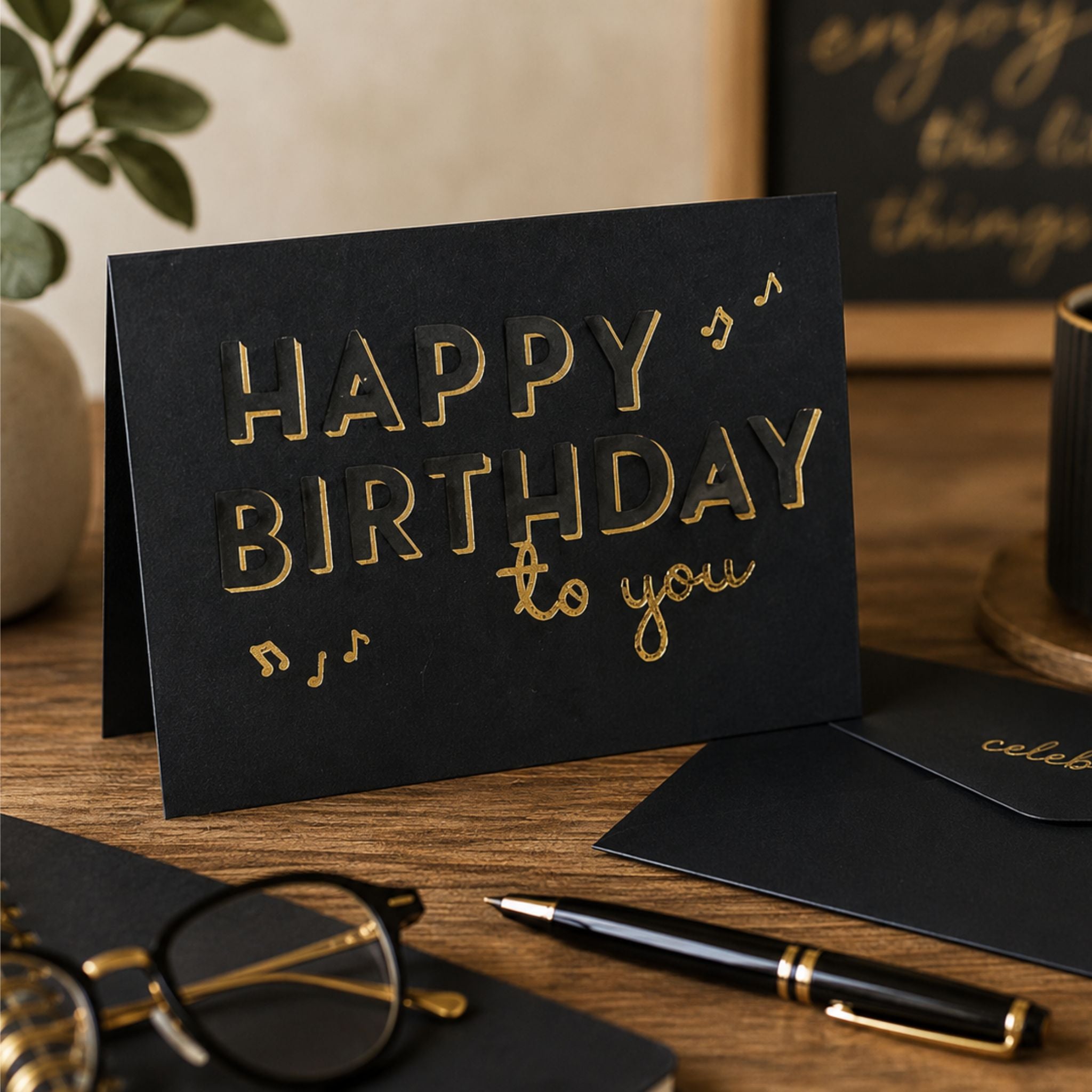 A black greeting card with gold foil lettering that reads 'HAPPY BIRTHDAY to you' stands open on a wooden desk, surrounded by a pen, eyeglasses.