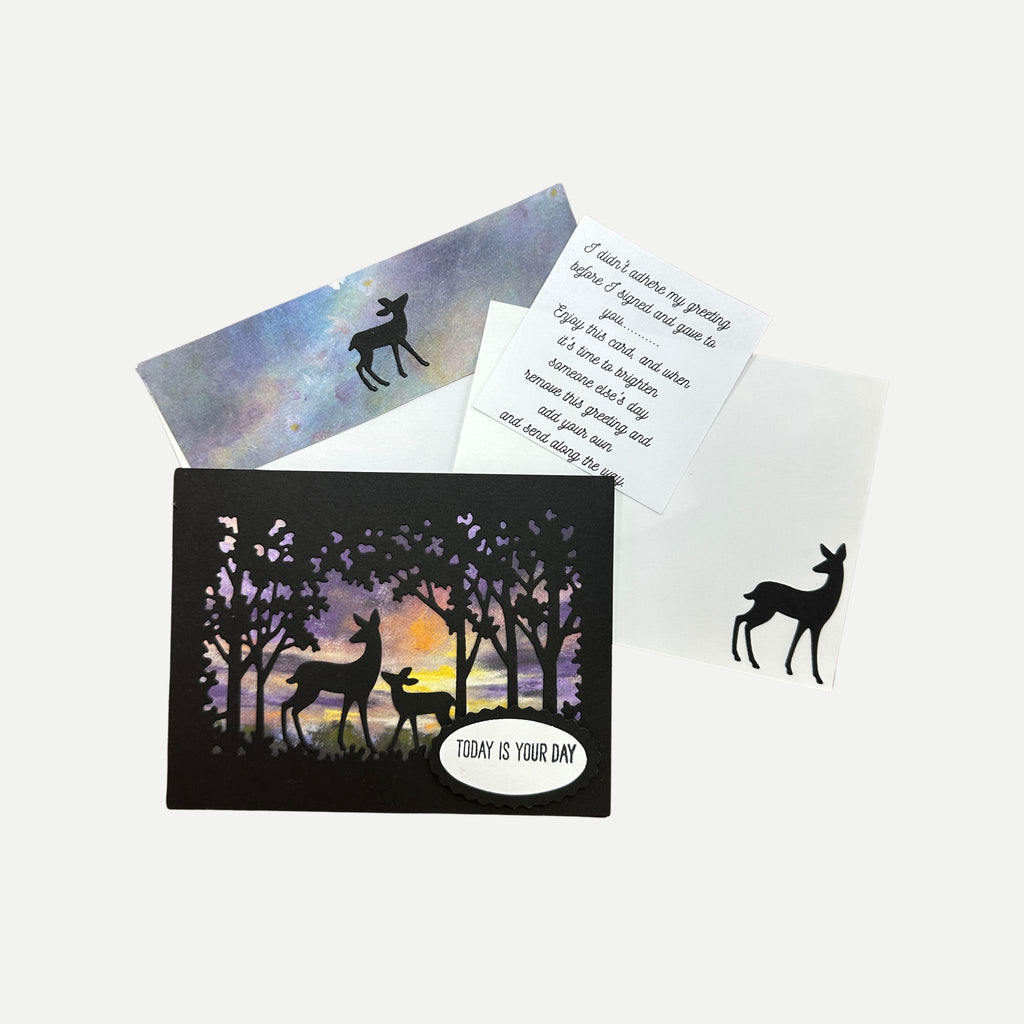 A greeting card set with a black silhouette forest and deer design, a white card with printed message text, and a blue-purple envelope on a light gray background.