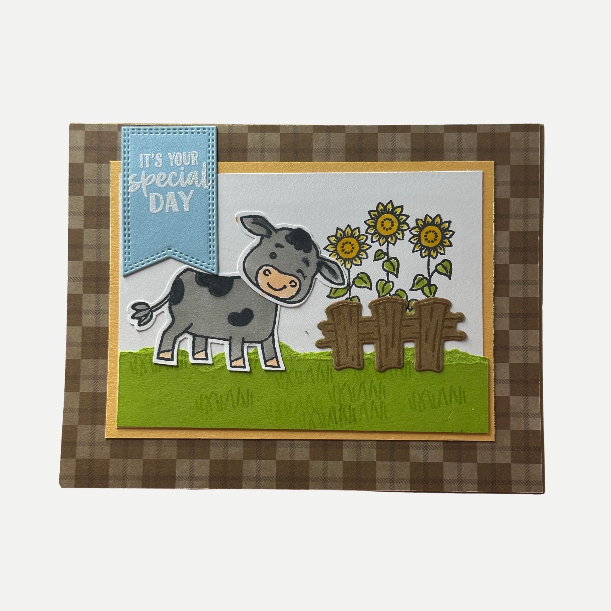 Handmade rustic birthday card featuring a smiling cow beside a sunflower fence with plaid background.
