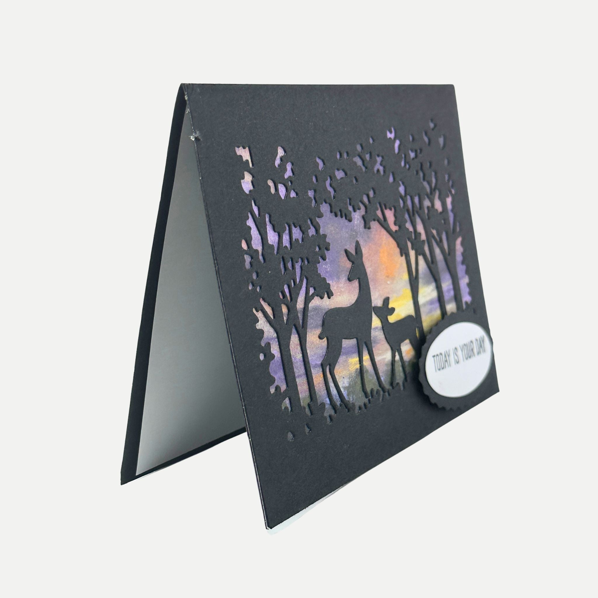 A black greeting card with a laser-cut forest silhouette design featuring deer and trees, set against a colorful sunset sky, with a small oval white sticker on the front.