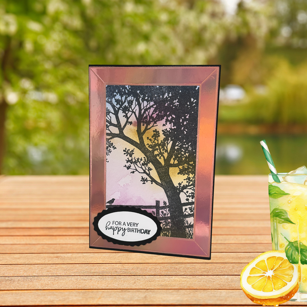 Handmade birthday card featuring a silhouetted tree against a soft pink and gold sunset, displayed outdoors on a wooden table with a refreshing lemonade beside it.